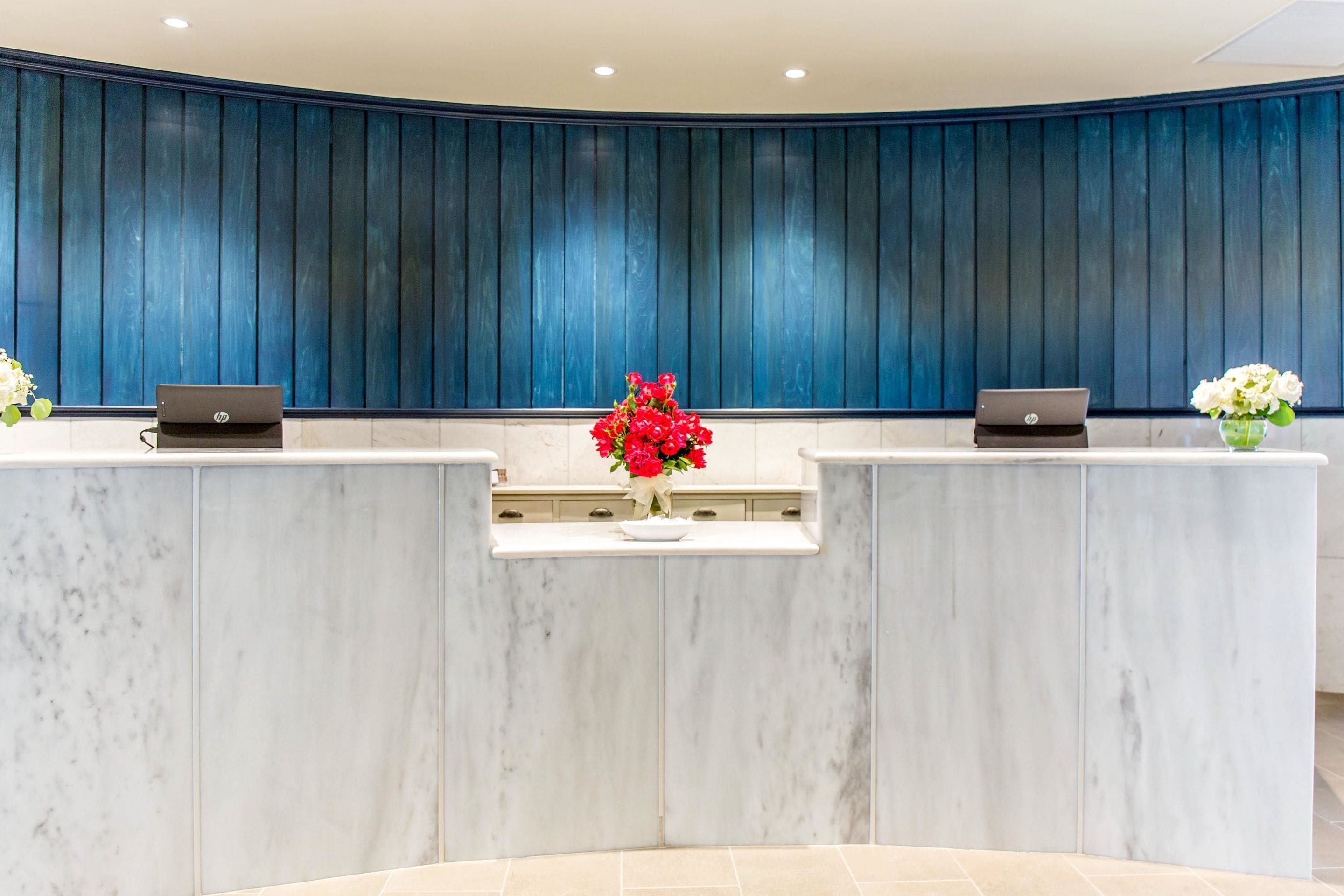 A reception desk with two laptops, a vase of red flowers, and a blue paneled wall.