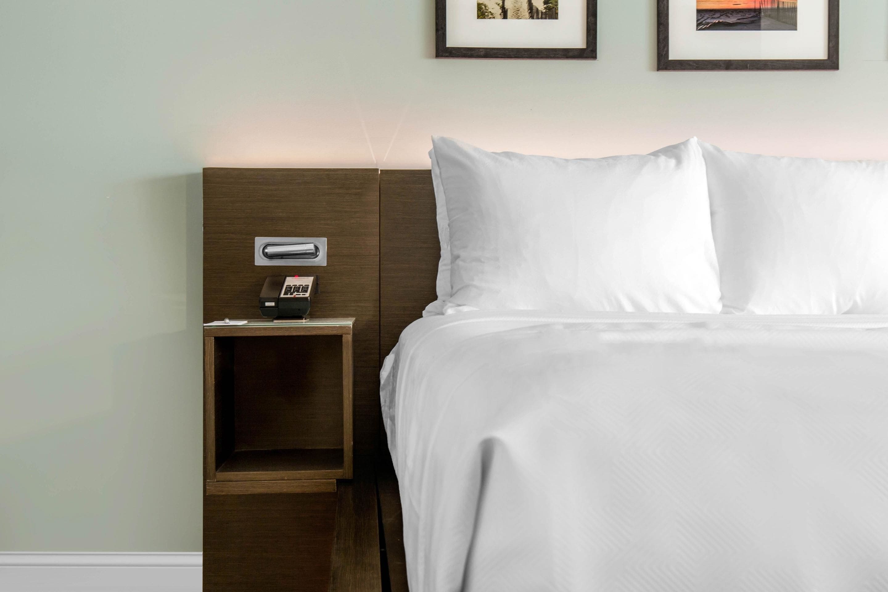 Cama bem arrumada com lençóis brancos, mesa de cabeceira de madeira com abajur e um quadro na parede.