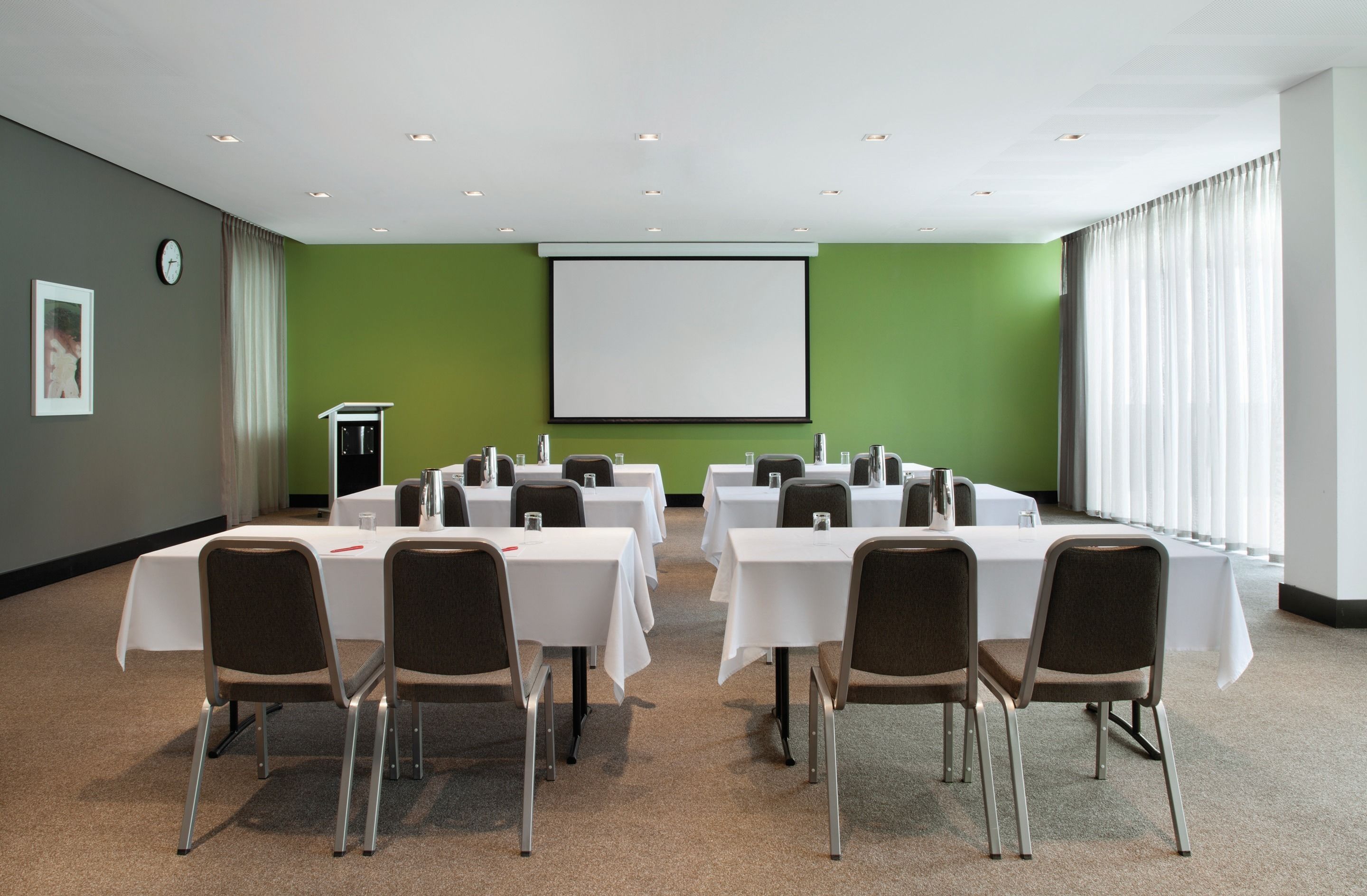 A conference room with multiple tables and chairs, a projector screen, and a clock on the wall.