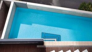 Una piscina rectangular con agua azul cristalina, rodeada por un borde de baldosas y una estructura de madera.