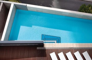 En rektangulær swimmingpool med klart blåt vand, omgivet af en flisbelagt terrasse og en trækonstruktion.