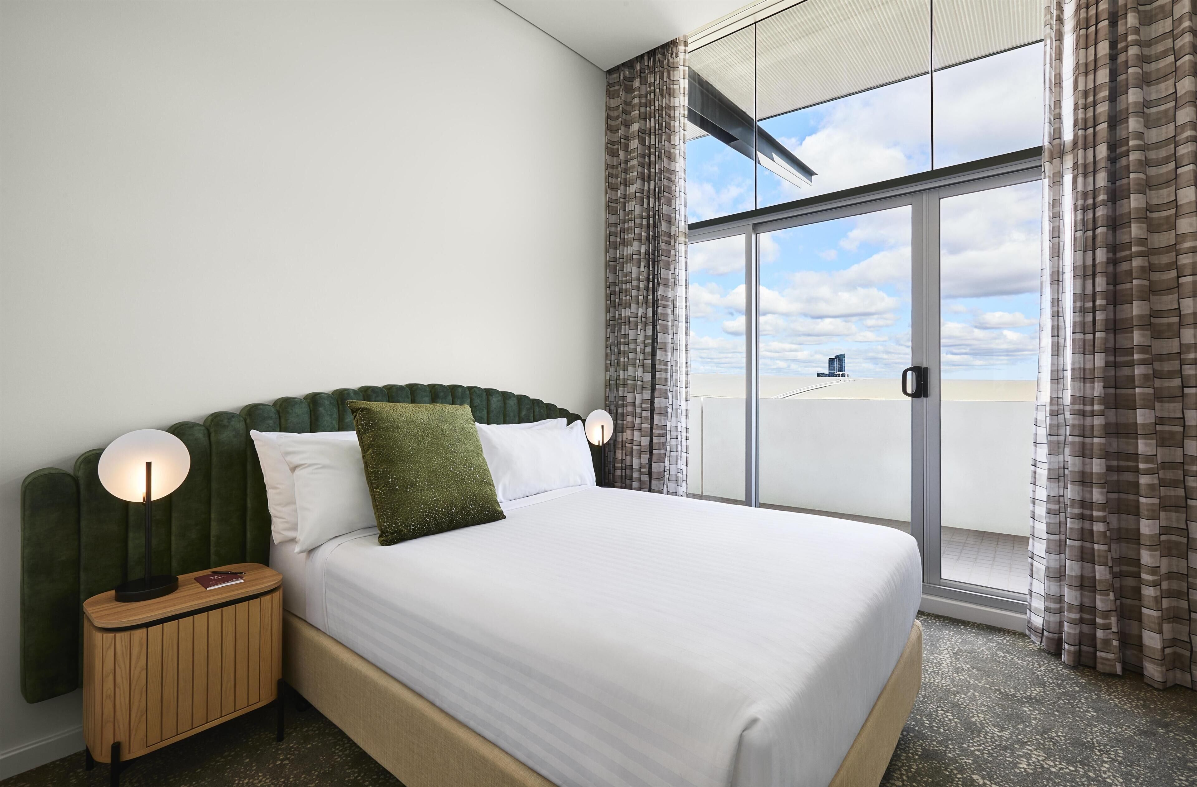 Habitación de hotel con cama, mesita de noche, lámpara y vistas al cielo y edificios.