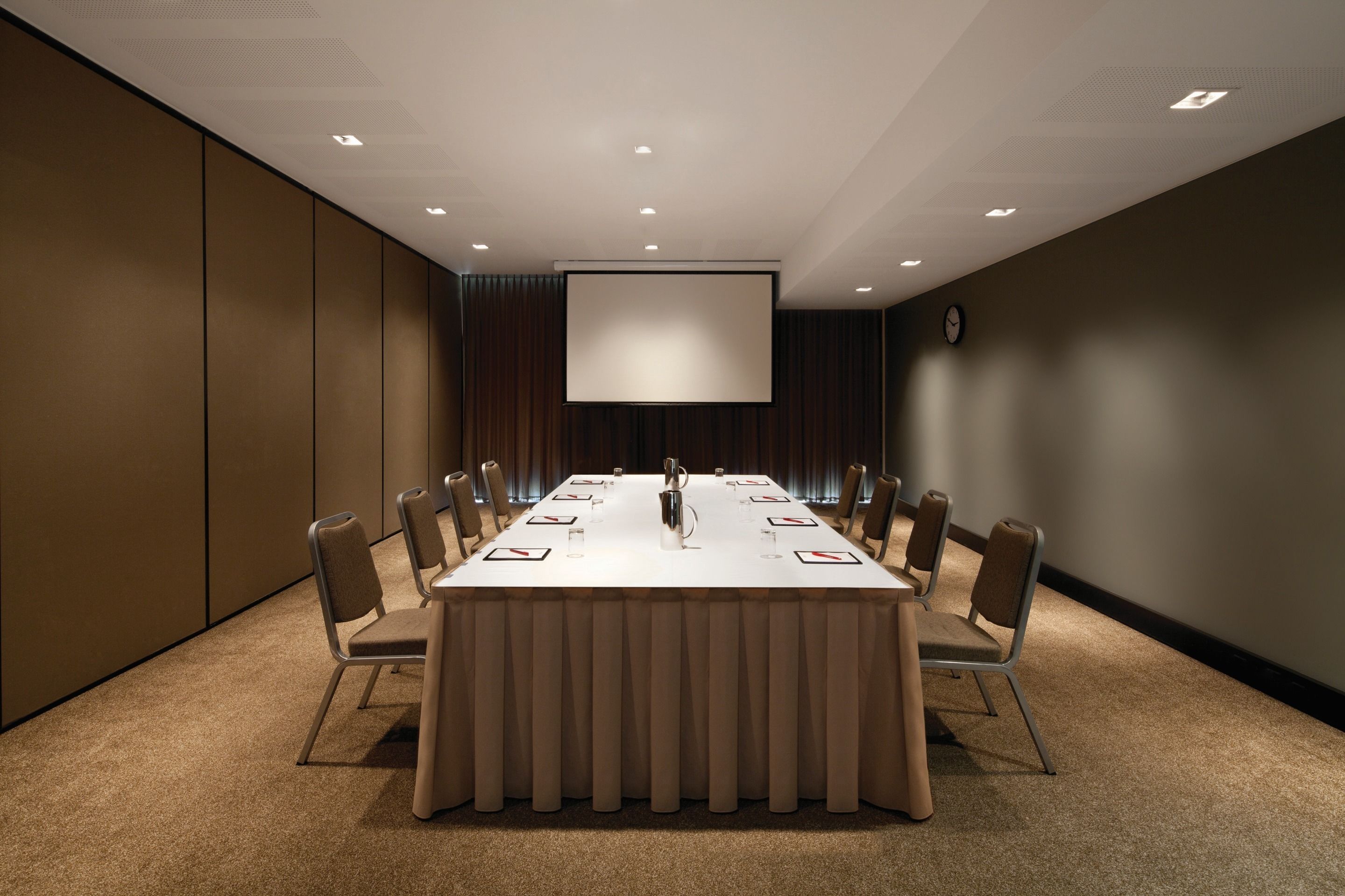 A conference room with a long table, chairs, a projector screen, and a clock on the wall.