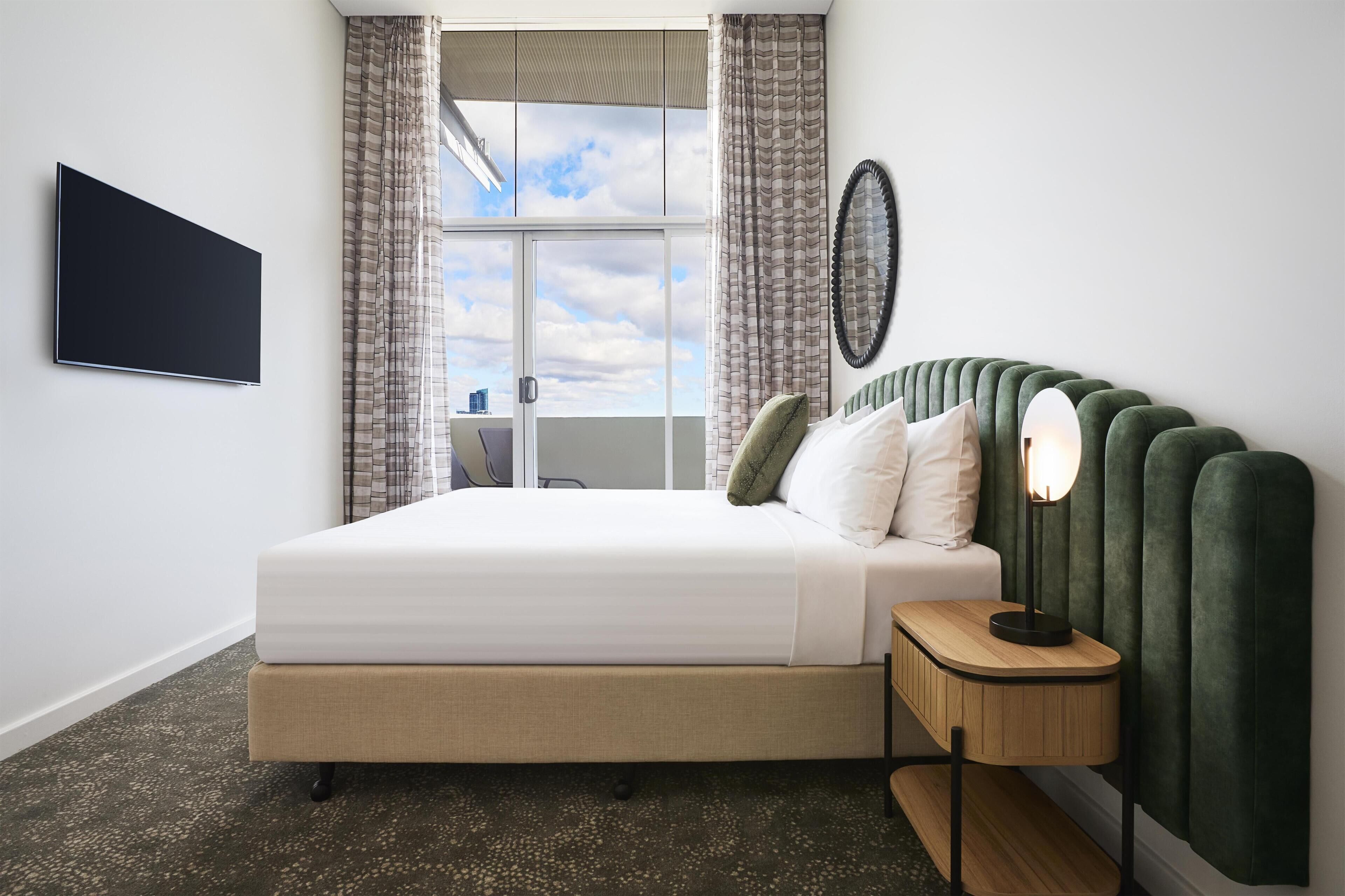A modern hotel room with a large bed, a flat-screen TV, a bedside table with a lamp, and a view of the sky through the window.