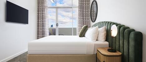 A modern hotel room with a large bed, a flat-screen TV, a bedside table with a lamp, and a view of the sky through the window.