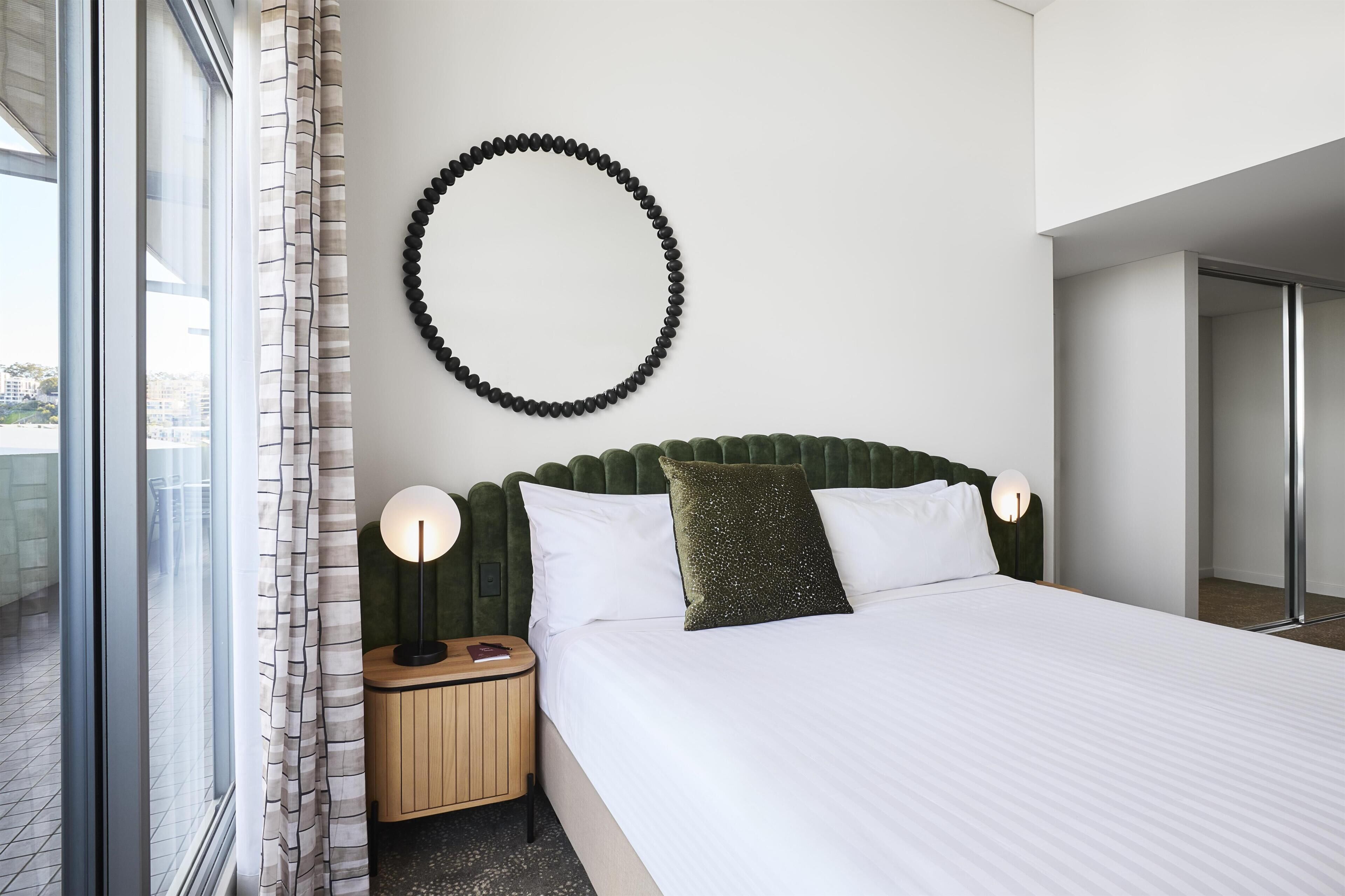 Ein modernes Hotelzimmer mit einem großen Bett, einer runden Wanddekoration, einem Nachttisch mit Lampe und Blick durch ein Fenster.