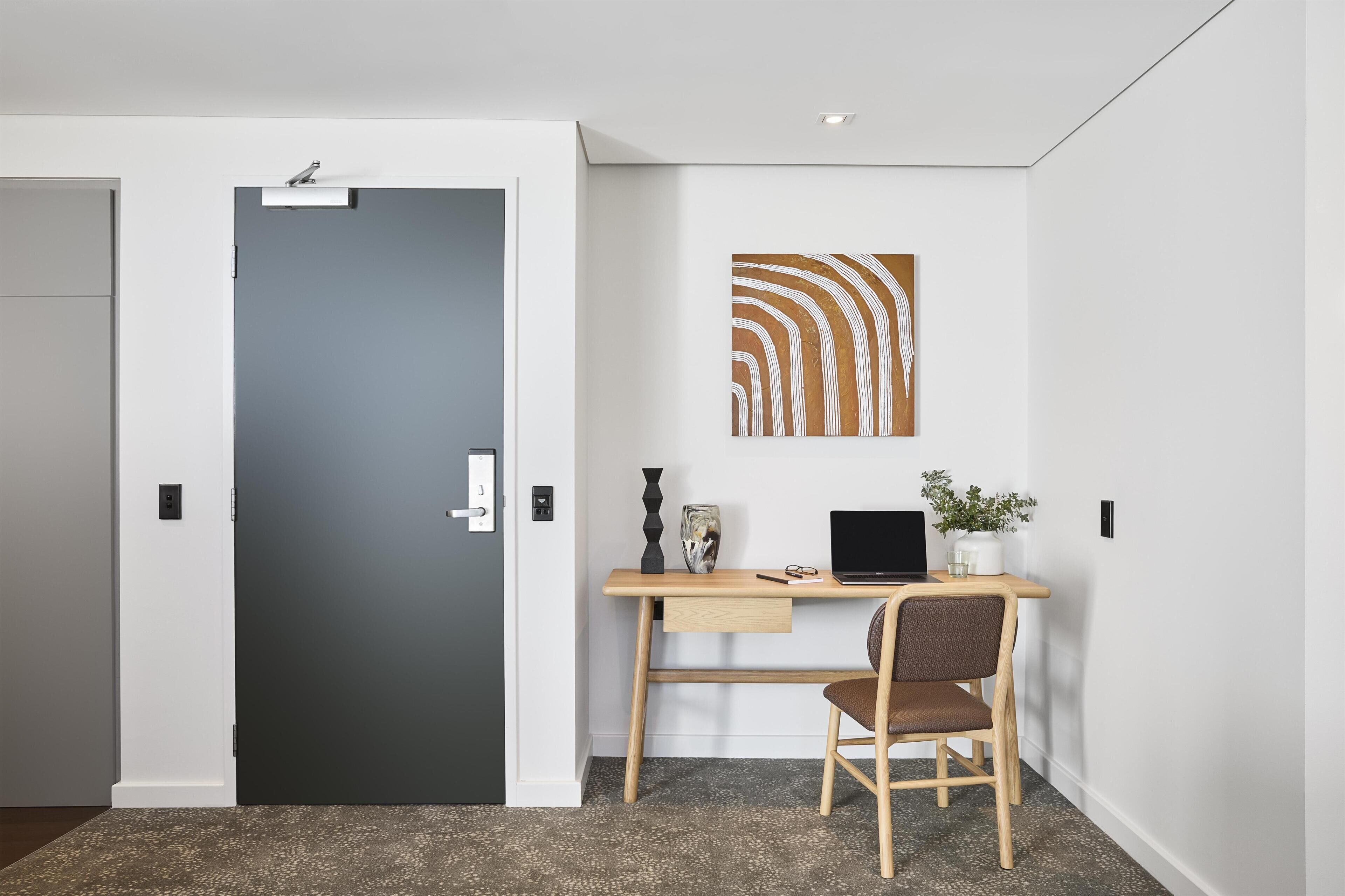 A modern office space with a dark door, a wooden desk, a laptop, a chair, and a wall-mounted abstract art piece.