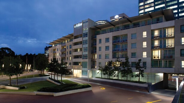 Un hotel moderno con un llamativo letrero de 'Adina', varios balcones y una zona de entrada bien iluminada.