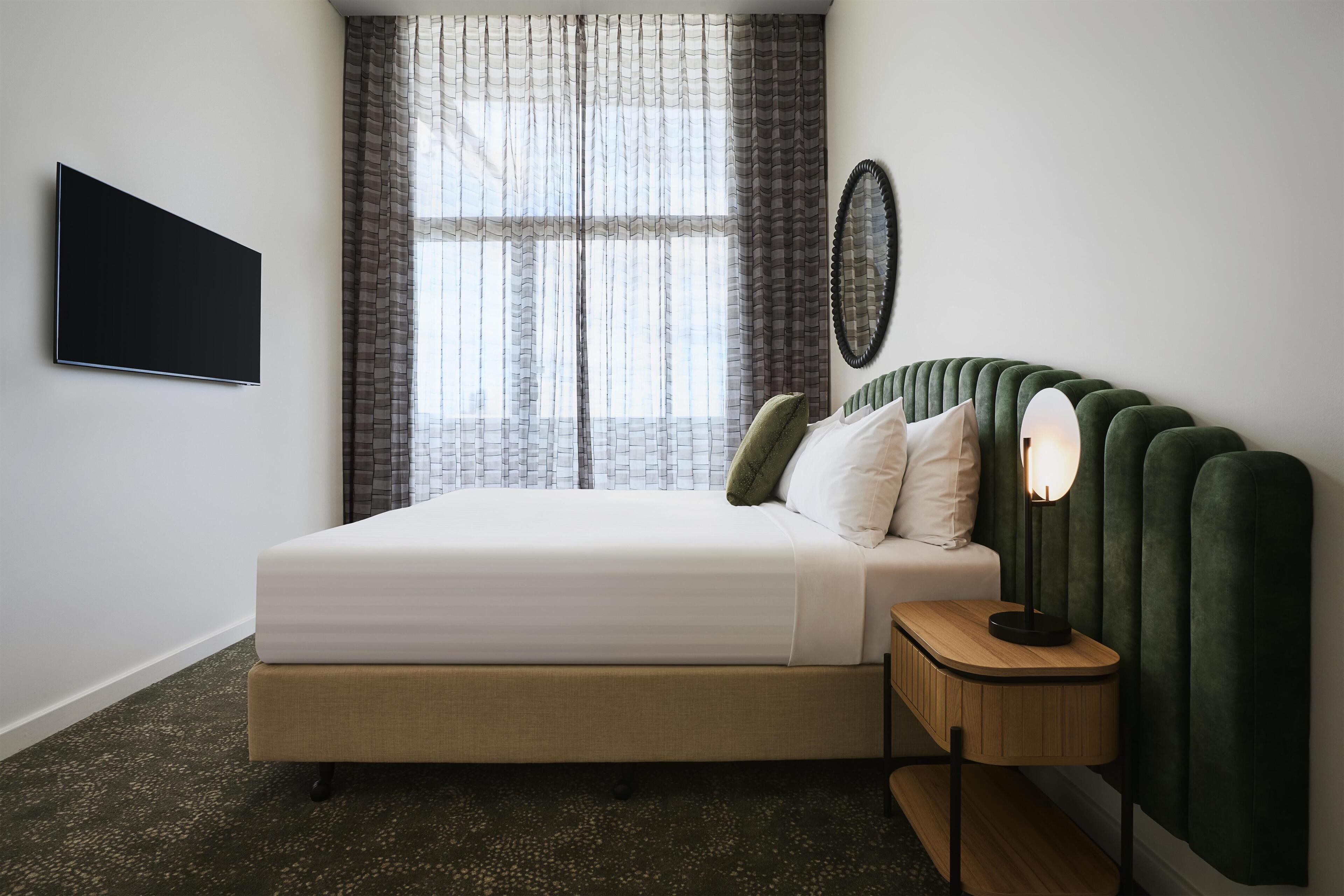 A modern bedroom with a bed, a bedside table with a lamp, a wall-mounted TV, and a window with curtains.