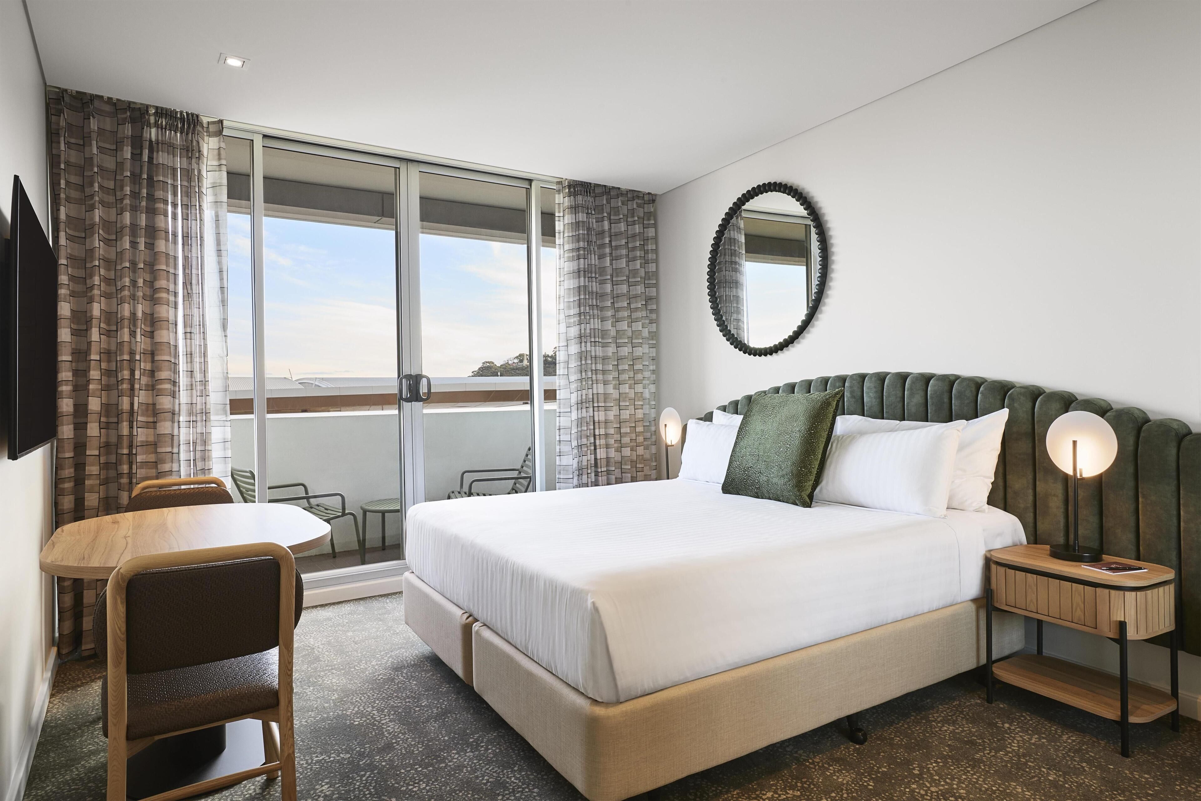 Ein Hotelzimmer mit einem großen Bett, einem Schreibtisch, einem Sessel und einem runden Spiegel, mit Blick durch die Glastüren.