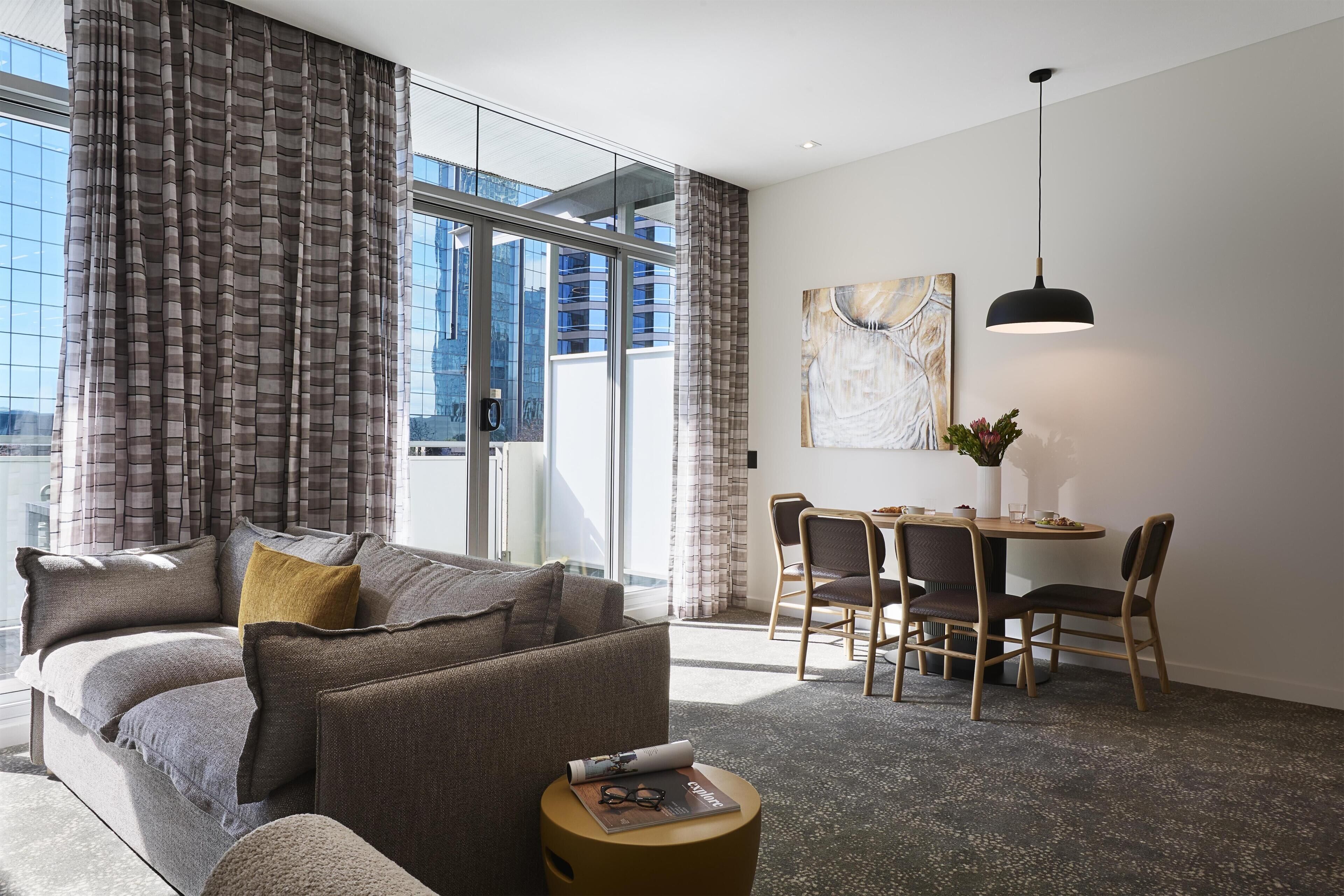 A modern living room with a sofa, a dining table with chairs, and a large window with curtains.