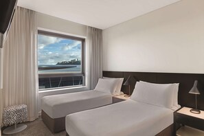 Two single beds with white linens, each with a nightstand and lamp, offer a serene view of a body of water.