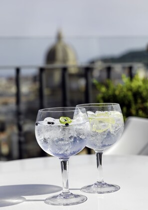 Deux verres contenant une boisson claire, avec des glaçons et une tranche de citron vert en guise de décoration, sont posés sur une table blanche, le tout sur fond de paysage urbain.