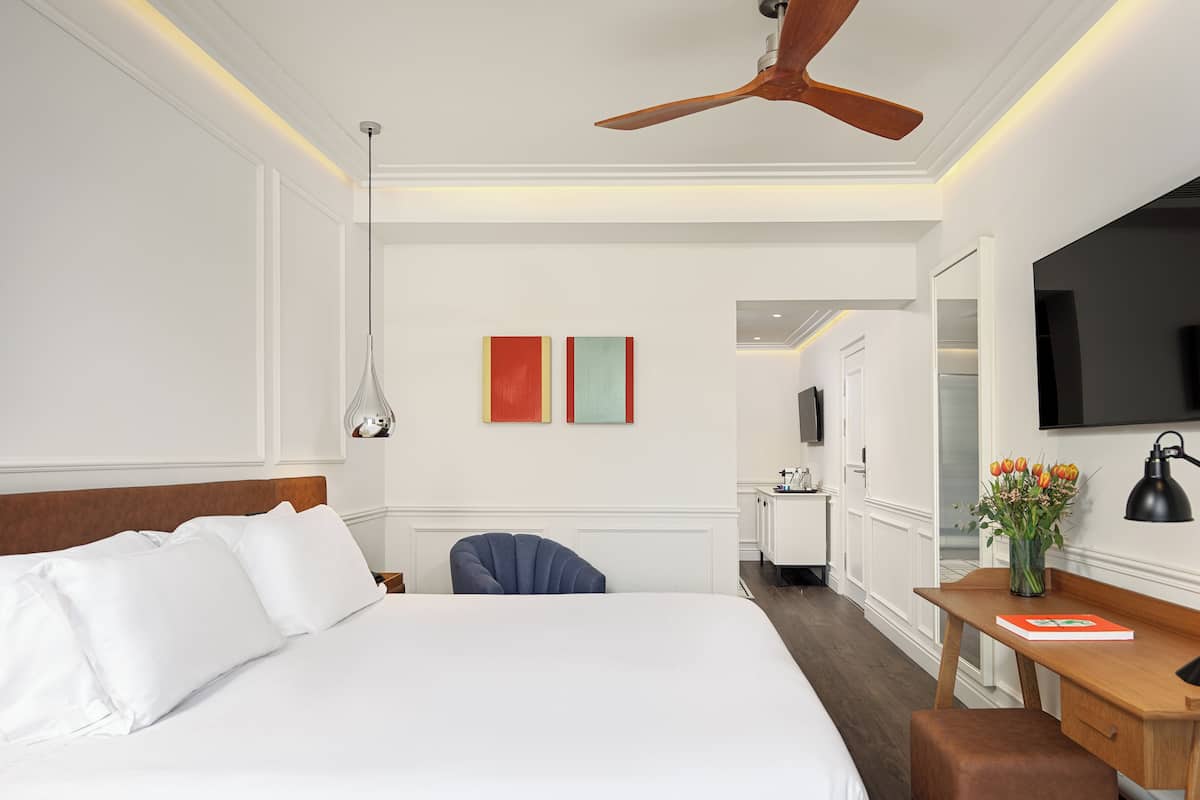 Een hotelkamer met een groot bed, een bureau met stoel, een plafondventilator, een televisie en een vaas met bloemen.