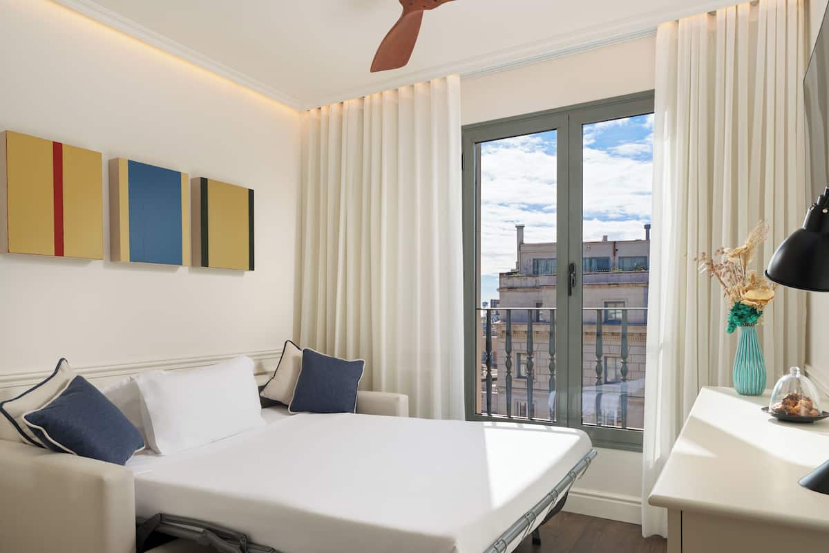 Een moderne hotelkamer met een bed, een bureau, een raam met gordijnen en uitzicht op gebouwen.