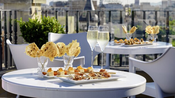 En tagterrasse med borde dækket til måltid, med et udvalg af appetizers og glas hvidvin, med udsigt over bybilledet.
