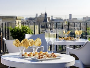 Une terrasse sur le toit, avec des tables dressées pour un repas, où l’on propose une variété d’amuse-gueules et des verres de vin blanc, et d’où l’on peut admirer la vue sur la ville.