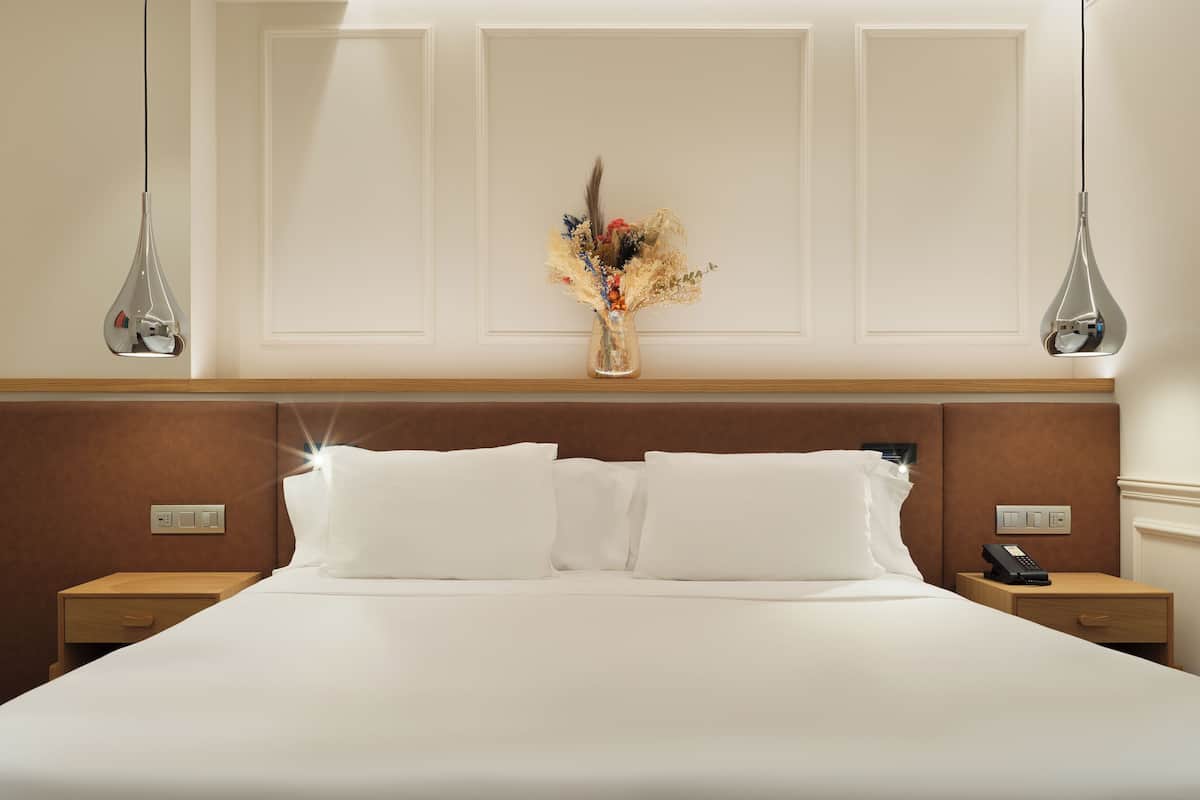 Hotelkamer met een groot bed, twee nachtkastjes, een bijzettafeltje met telefoon, een vaas met bloemen en hanglampen.