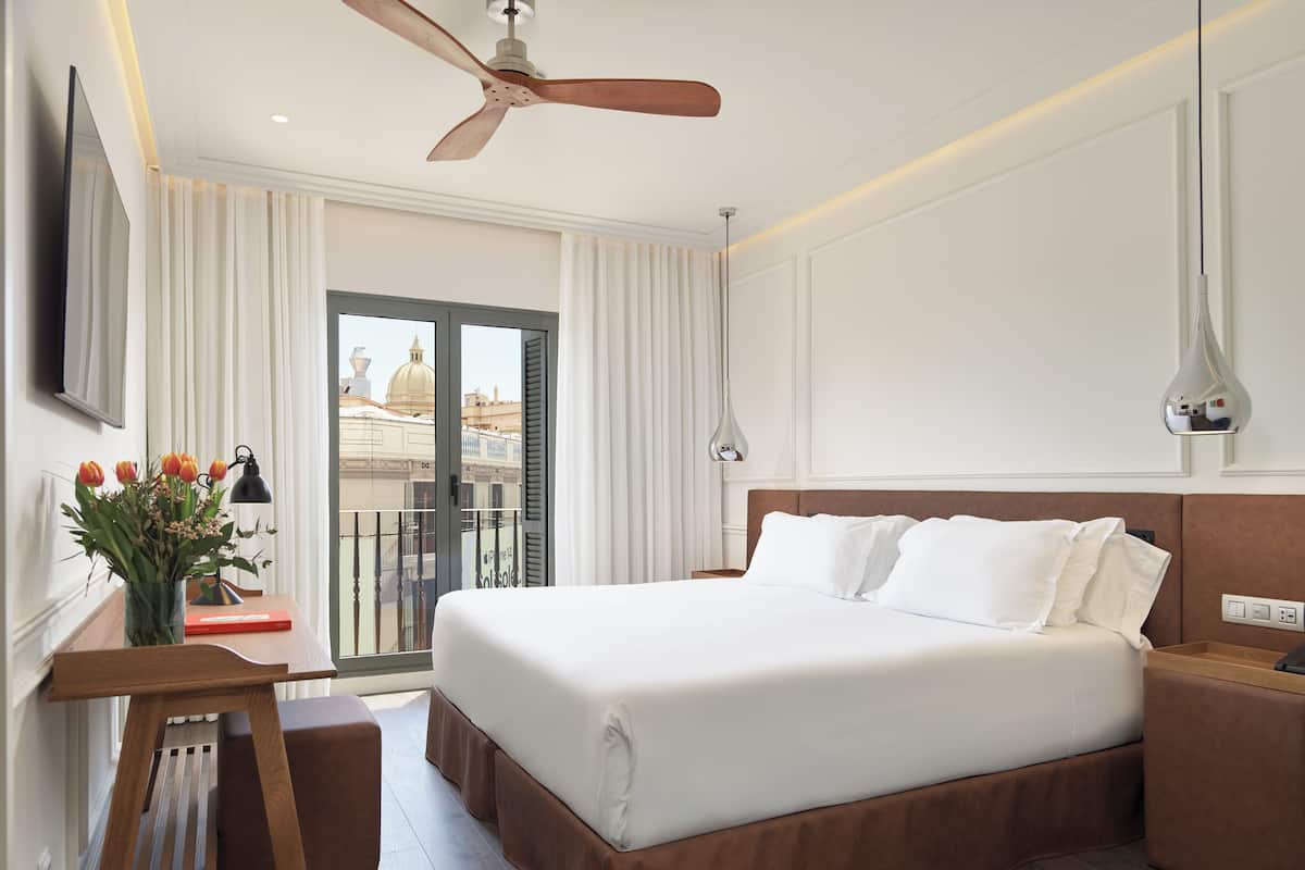 Een hotelkamer met een groot bed, een bureau met lamp, een bloevas en uitzicht op gebouwen.