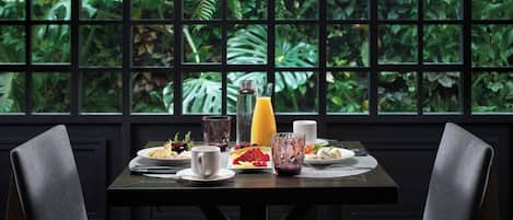 Mesa lista con platos, vasos y una cesta de frutas, con vistas a un exuberante follaje desde la ventana.