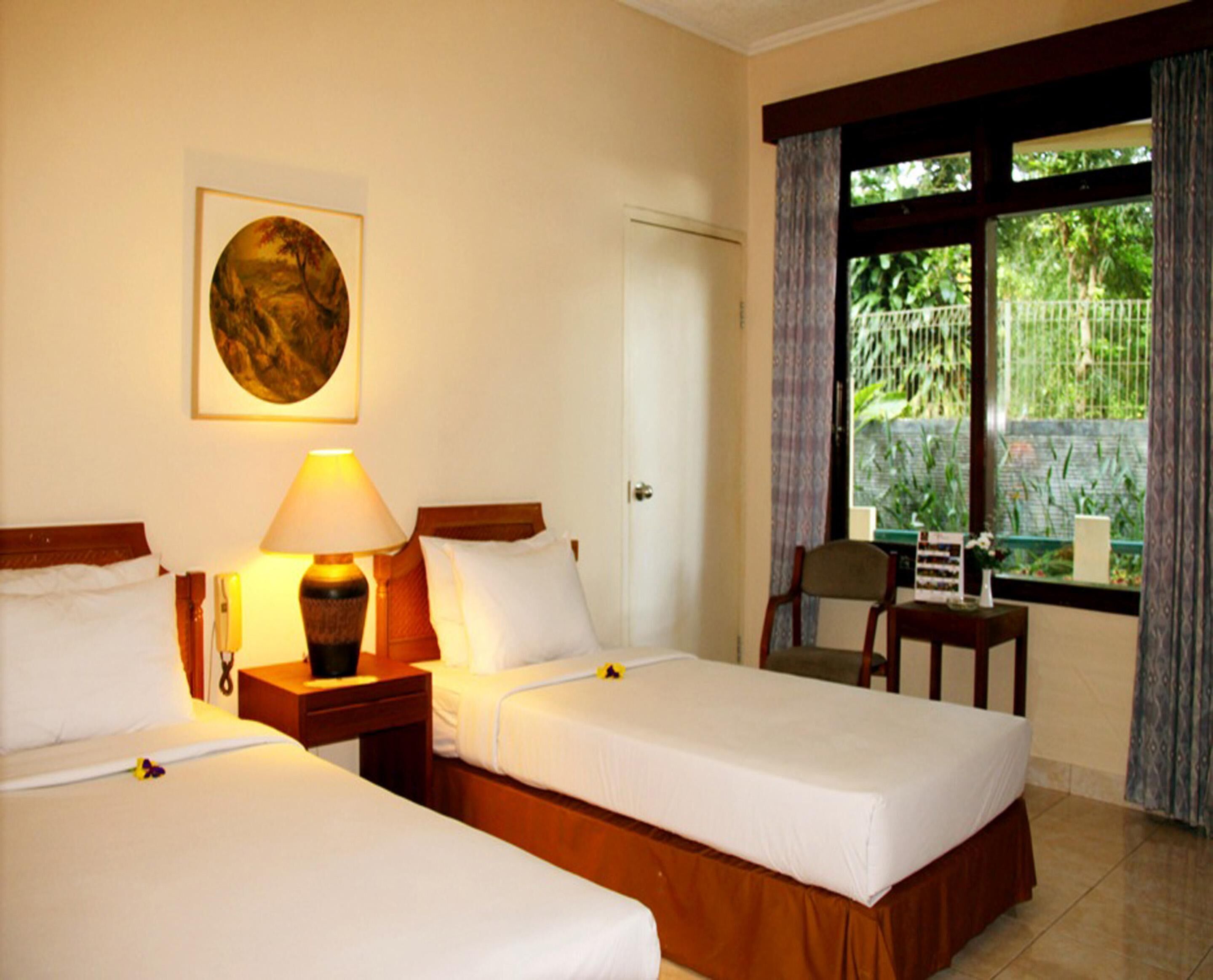 A hotel room with two single beds, a bedside lamp, a framed picture on the wall, a window with a view of greenery, and a small table with a chair.