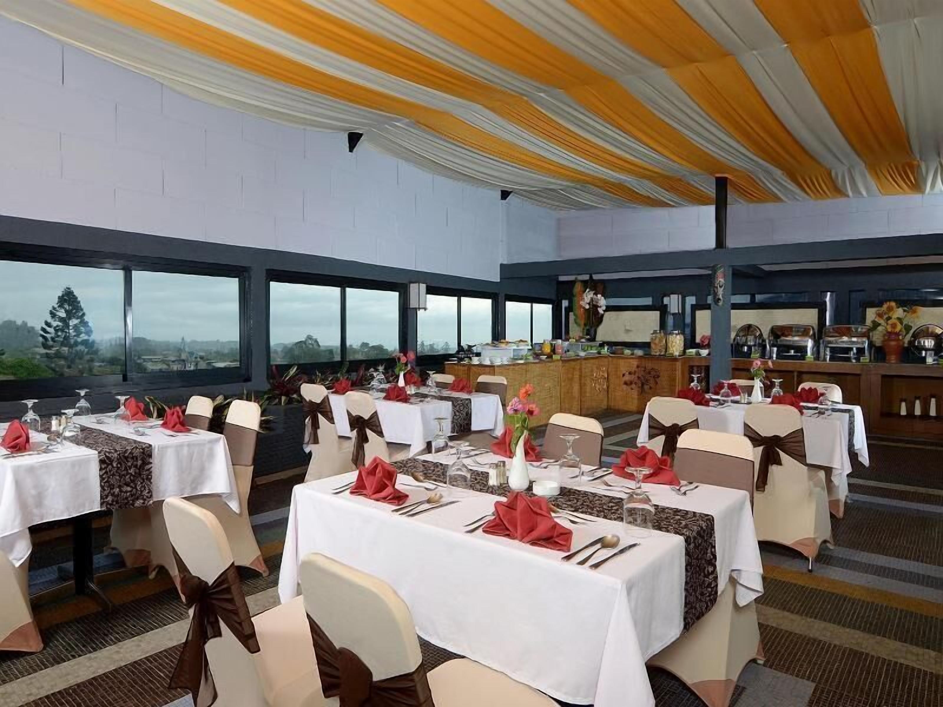 A banquet hall with multiple round tables, each set with white tablecloths and red napkins. The room has large windows offering a view of the outdoors.