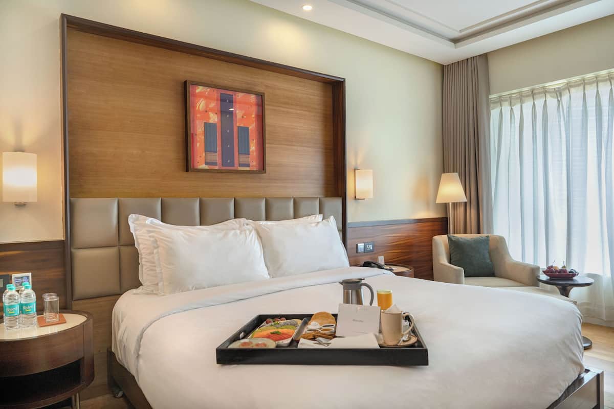 Una habitación de hotel con una cama grande, un cabecero con una obra de arte enmarcada, una mesita de noche con una lámpara, una silla y una bandeja con alimentos para el desayuno.