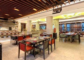 A modern restaurant with multiple dining areas, a kitchen in the background, and a ceiling with wooden slats and recessed lighting.