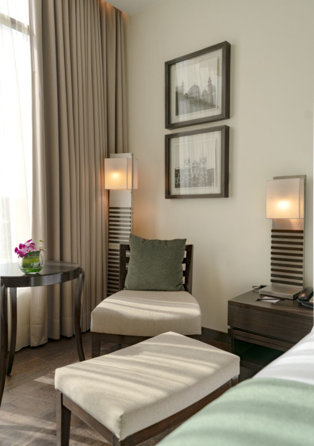 A hotel room with a bed, a chair, a small table with a vase of flowers, and two wall-mounted lamps.