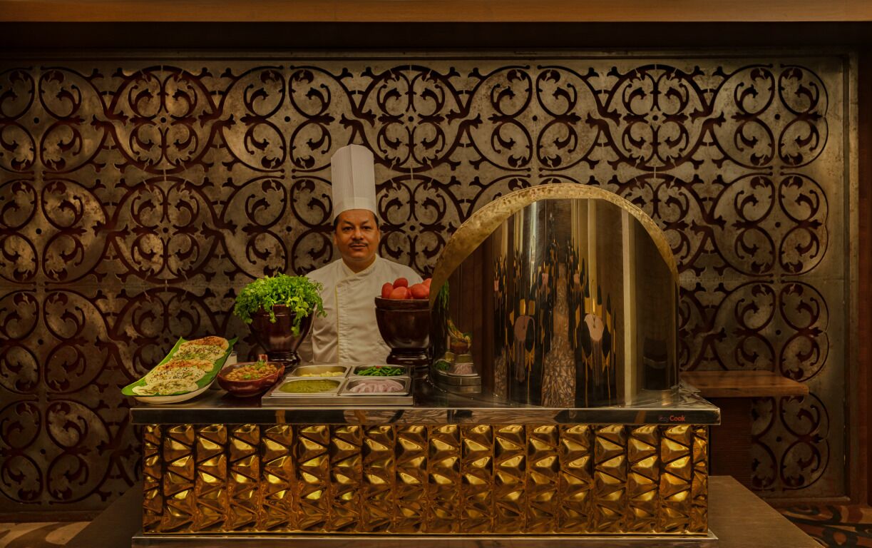 A chef standing behind a counter with various food items and a decorative mirror.