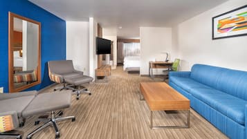 A hotel room with a blue sofa, a grey armchair, a wooden coffee table, a desk with a chair, a television, and a large mirror.