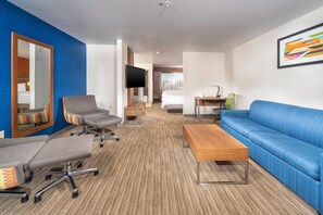 A hotel room with a blue sofa, a grey armchair, a wooden coffee table, a desk with a chair, a television, and a large mirror.