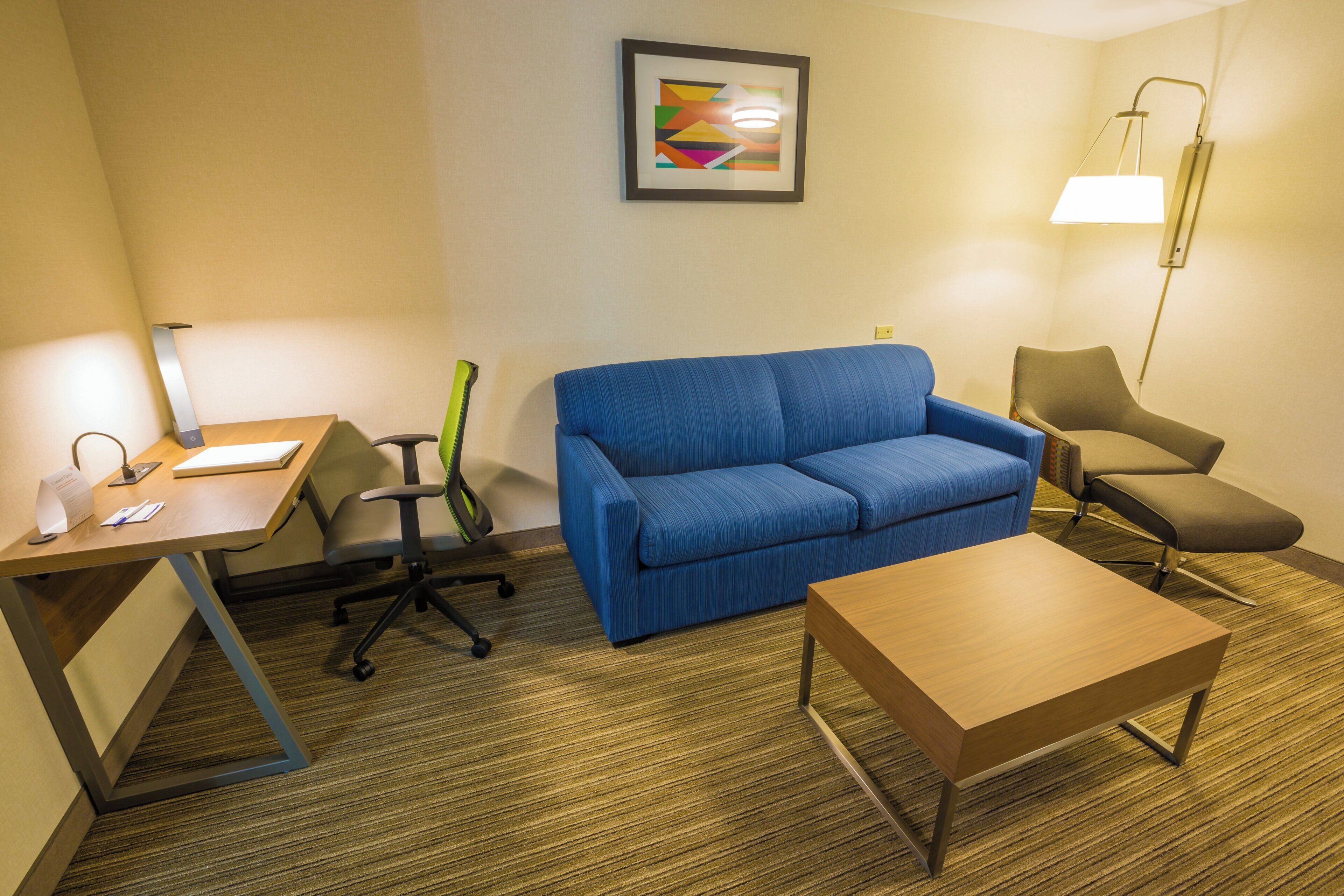 A hotel room with a desk, a blue sofa, a small table, a lamp, and a framed picture on the wall.