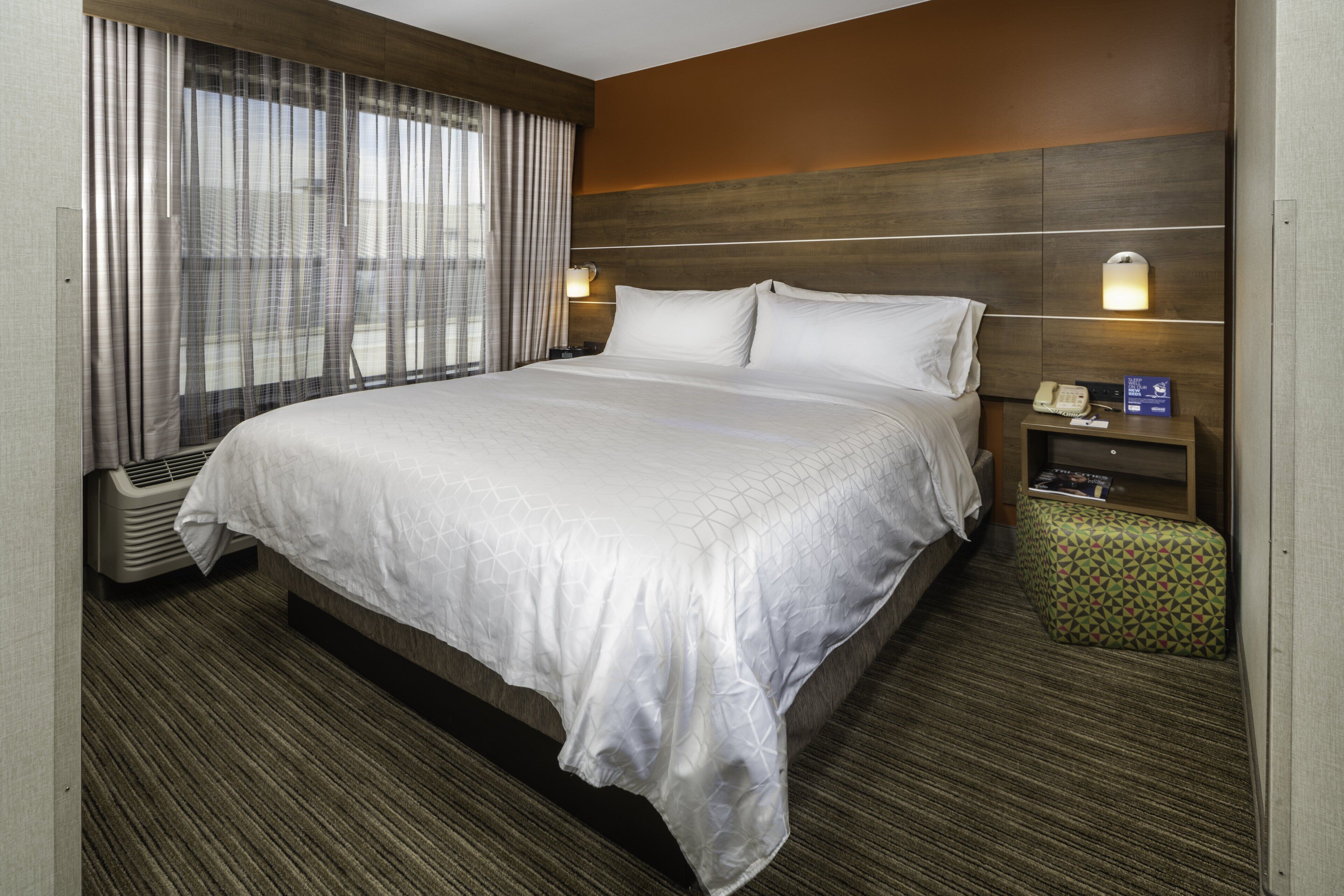 Habitación de hotel con una cama grande, una mesita de noche con un teléfono y una lámpara, un pequeño taburete verde y una ventana con cortinas.