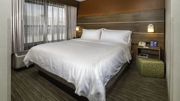 Habitación de hotel con una cama grande, una mesita de noche con un teléfono y una lámpara, un pequeño taburete verde y una ventana con cortinas.