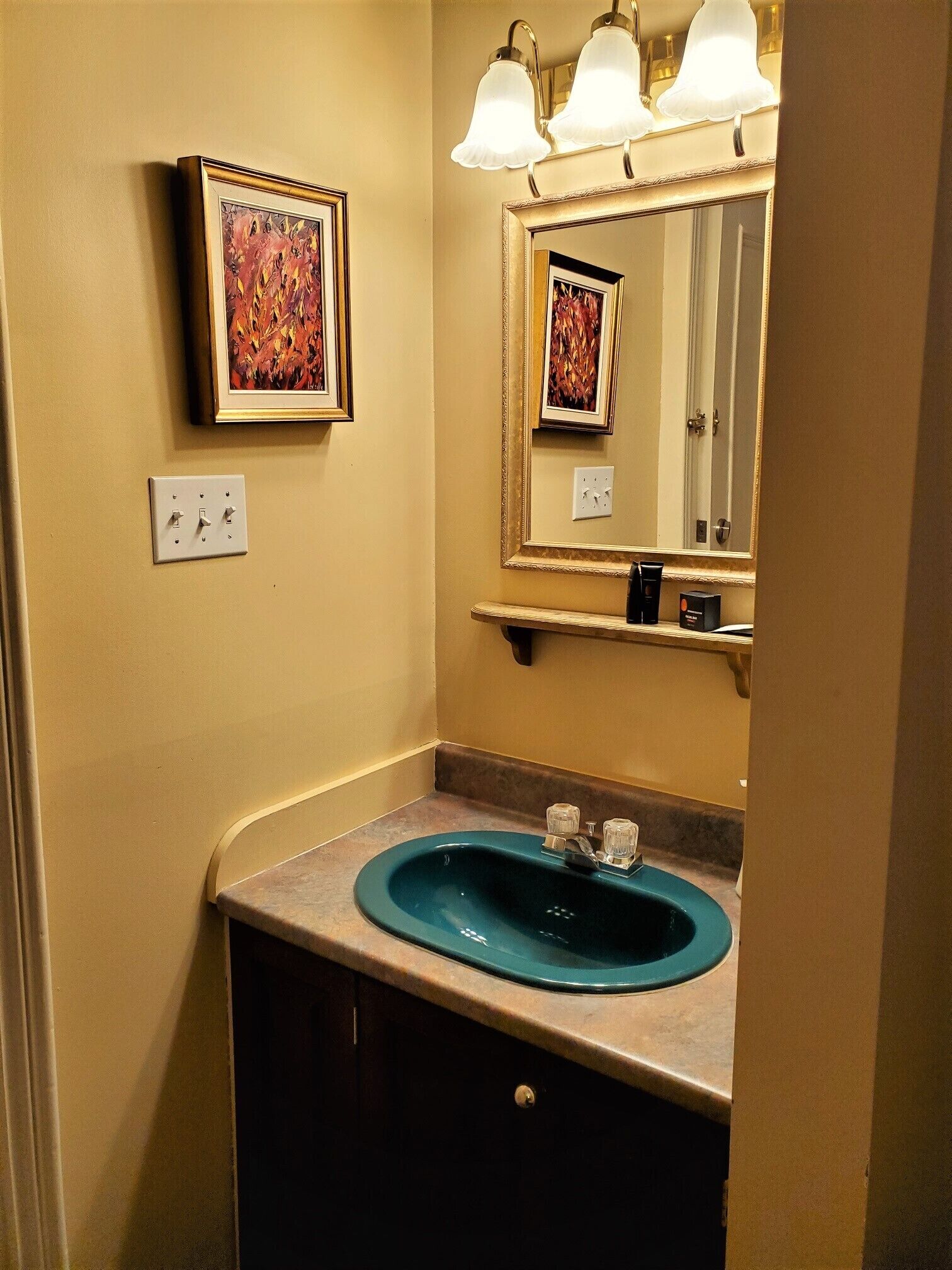 A bathroom with a blue sink, a mirror with lights, and a framed picture on the wall.