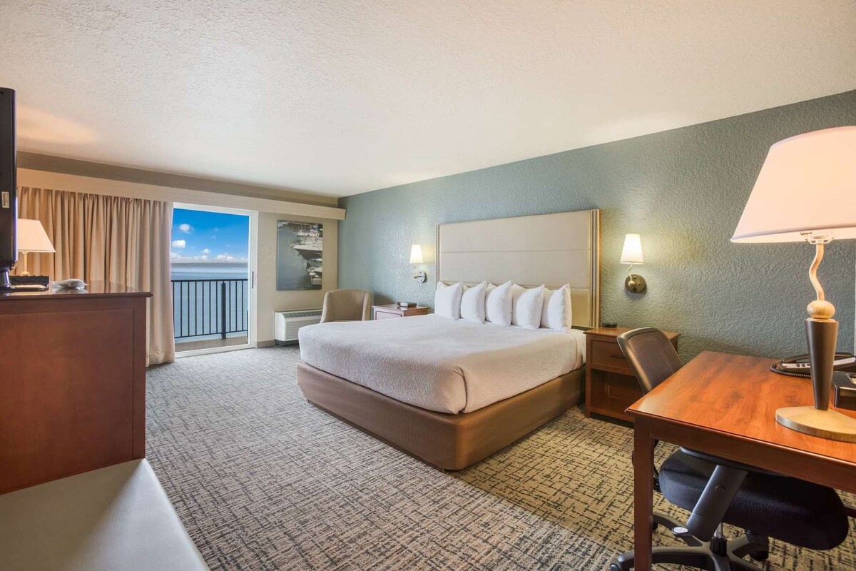 A hotel room with a bed, desk, chair, and a view of the sea.