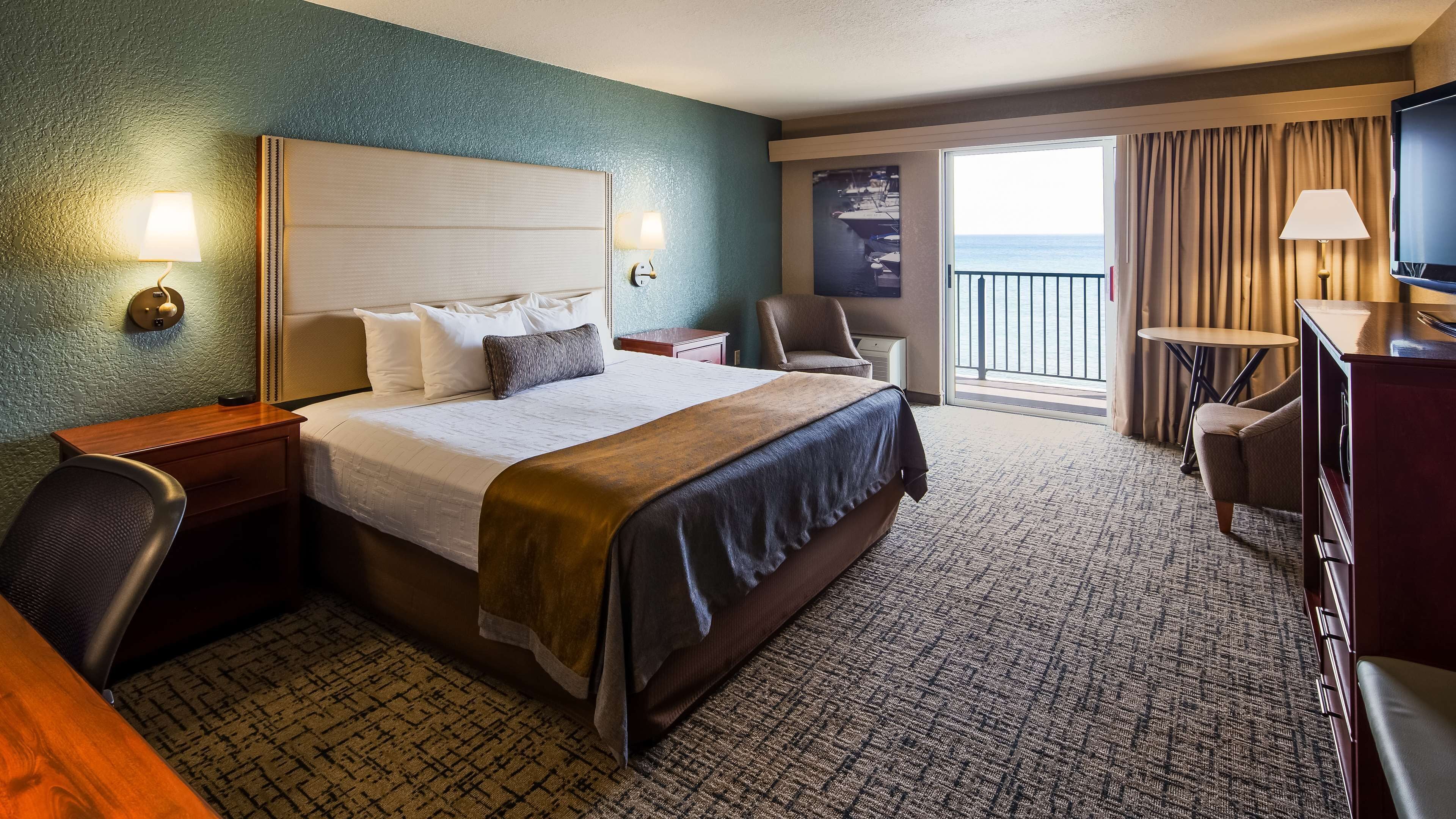 A hotel room with a large bed, a desk, a chair, a TV, and a balcony with a view of the sea.