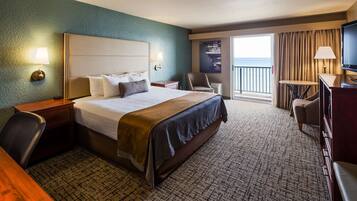 A hotel room with a large bed, a desk, a chair, a TV, and a balcony with a view of the sea.