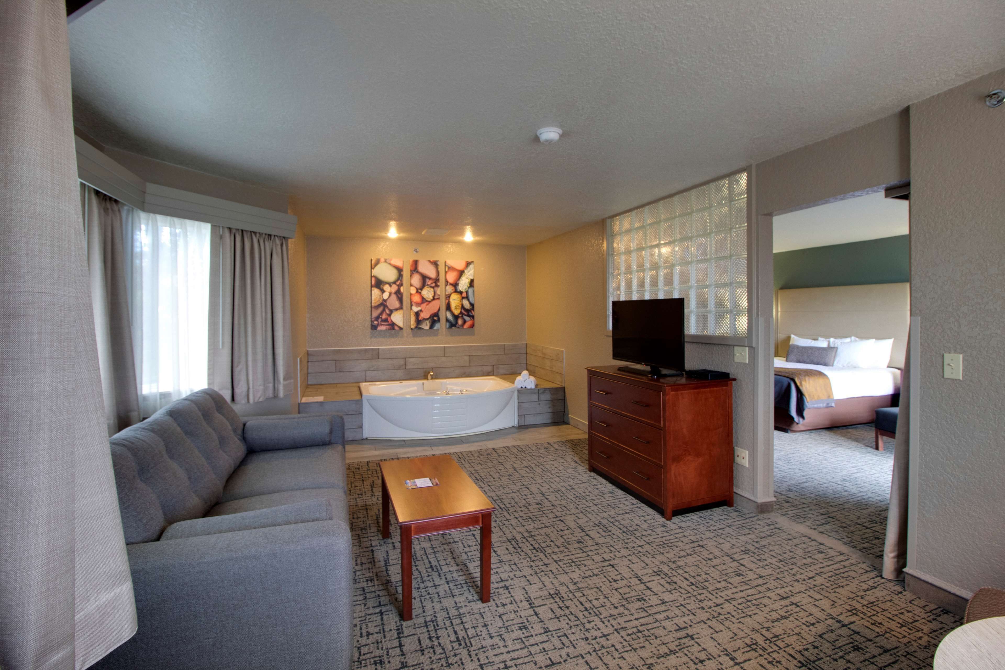 A modern hotel room with a sofa, a wooden table, a TV, a bathtub, and a painting on the wall.