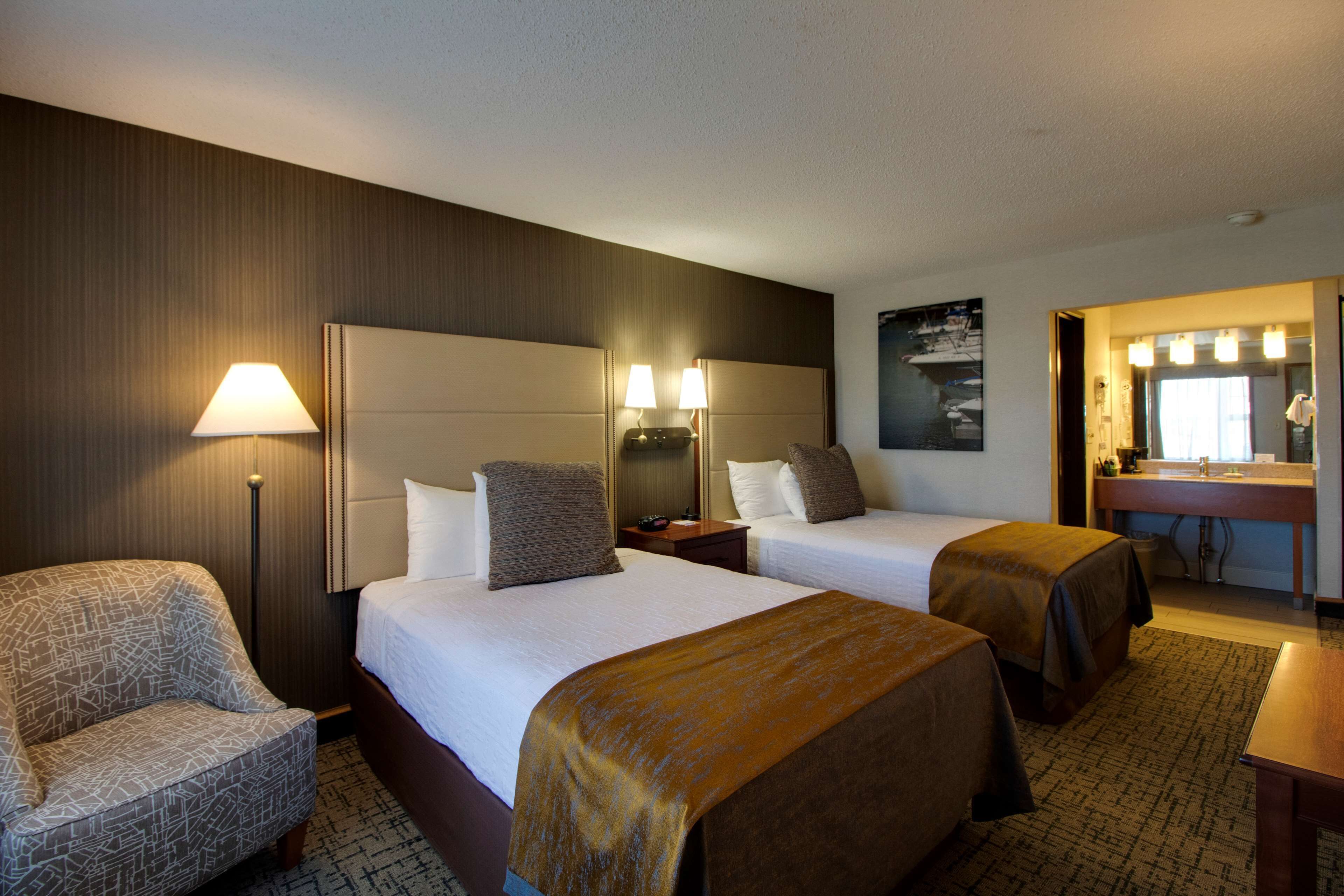 A hotel room with two beds, a chair, a desk, a mirror, and a bathroom with a sink and mirror.