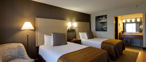 A hotel room with two beds, a chair, a desk, a mirror, and a bathroom with a sink and mirror.