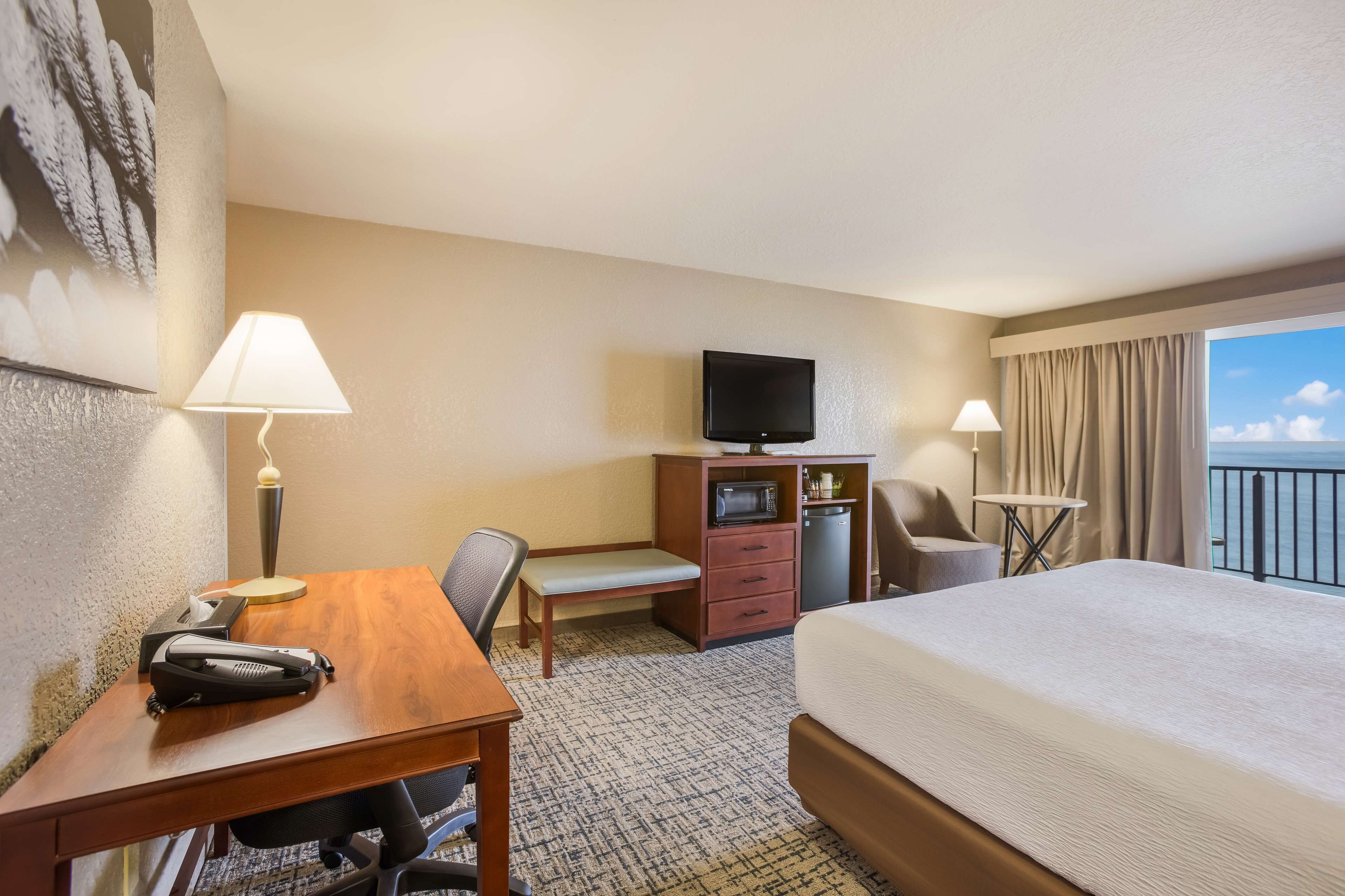 A hotel room with a bed, desk, chair, television, and a view of the sea.