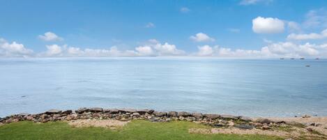 Un paysage côtier avec une zone herbeuse, un mur de pierre et une mer calme sous un ciel dégagé.