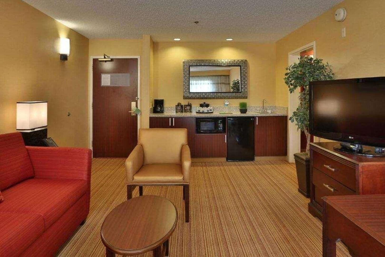 A hotel room with a red sofa, a beige armchair, a small wooden table, a TV, a kitchen area with a microwave, refrigerator, and a sink.