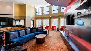 A spacious lobby with a blue sectional sofa, red chairs, a round coffee table, and a TV mounted on a red wall.
