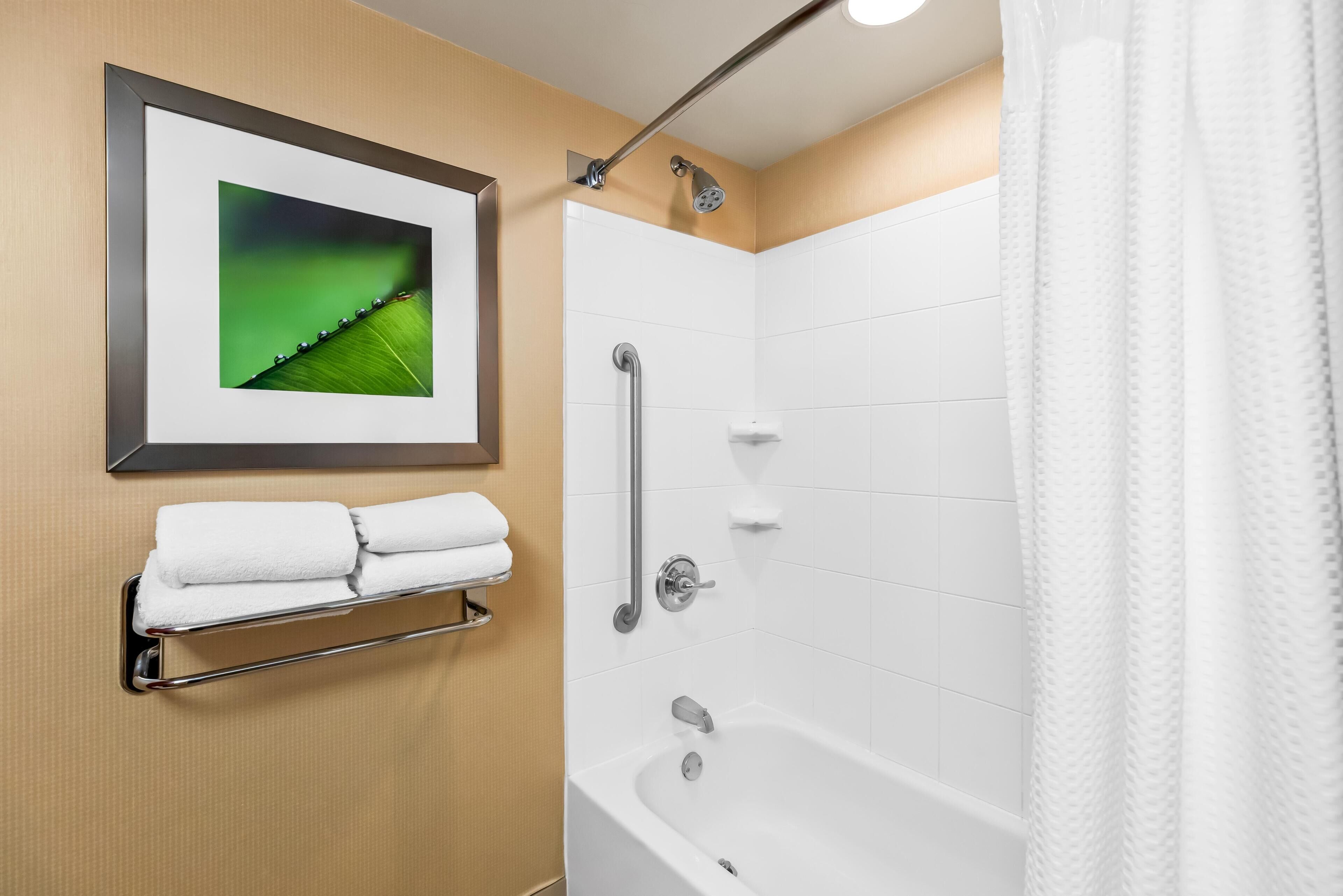 Een badkamer met een witte badkuip, een douche met een handdouche, een lijst met een foto aan de muur en een handdoekrek met opgevouwen handdoeken.