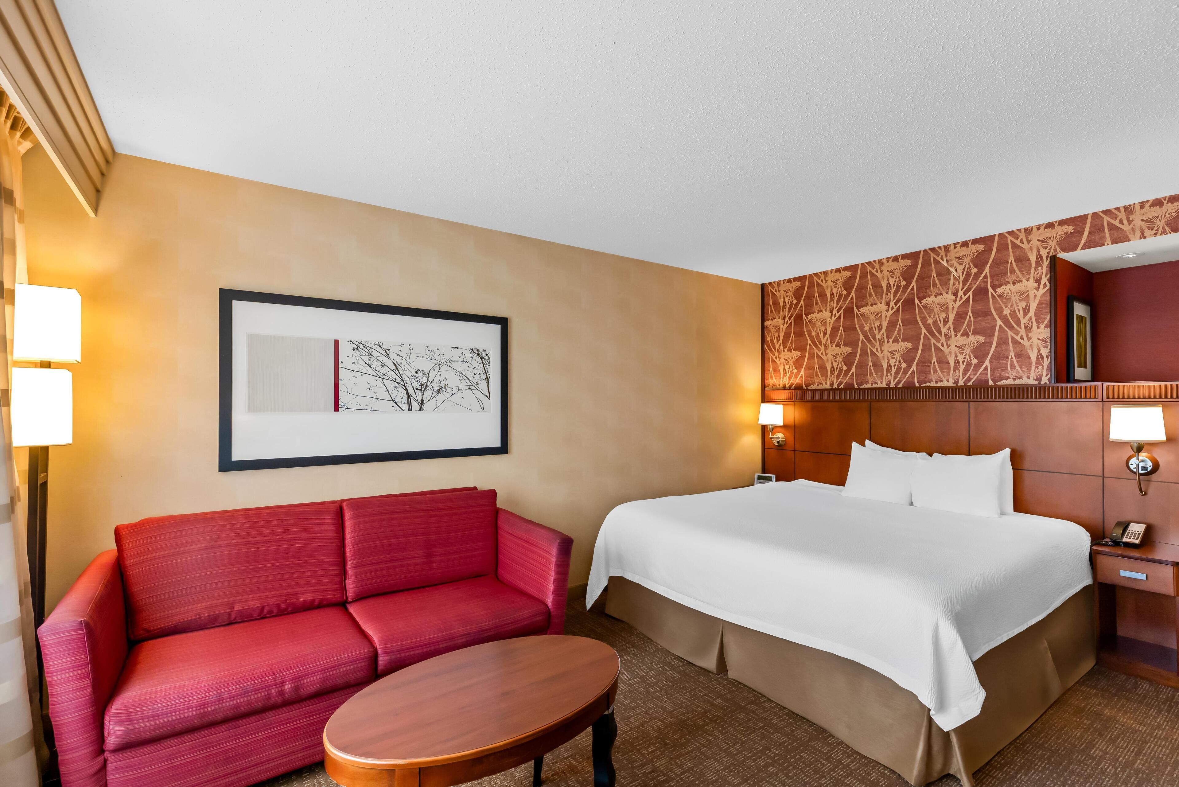 Habitación de hotel con un sofá rojo, una cama con ropa blanca, un cuadro en la pared y un papel tapiz estampado en la pared contigua.