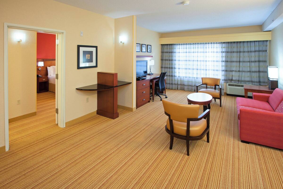 Habitación de hotel con sala de estar, escritorio, mesa de comedor con sillas, sofá rojo y dormitorio independiente con cama y mesita de noche.