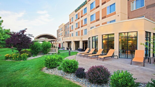 A hotel with a modern exterior, a covered outdoor seating area with lounge chairs, and a well-maintained garden. - Courtyard by Marriott Louisville Northeast (Louisville)